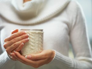 Woman holds a winter cup close up on light background. Woman hands with elegant french manicure nails design holding a cozy knitted mug. Winter and Christmas time concept.
